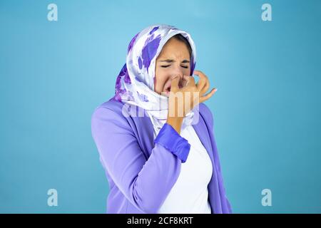 Jeune belle femme arabe portant le hijab islamique sur un fond bleu isolé qui sent quelque chose de stinky et dégoûtant, une odeur intolérable, tenant br Banque D'Images