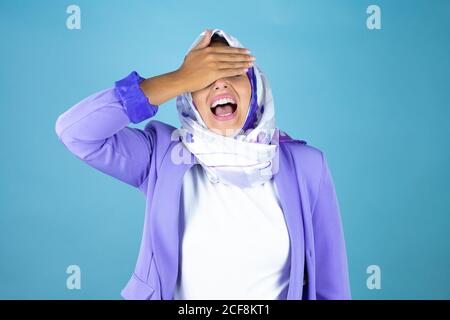 Jeune belle femme arabe portant le hijab islamique sur un bleu isolé arrière-plan sérieux et couvrant ses yeux avec sa main Banque D'Images