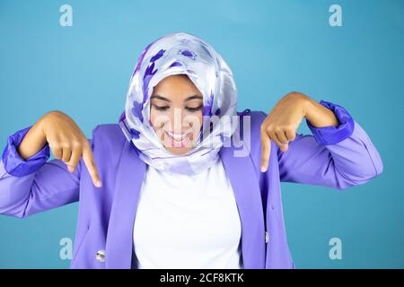 Jeune belle femme arabe portant le hijab islamique sur fond bleu isolé surpris, regardant la caméra et pointant vers le bas avec les doigts et la rai Banque D'Images