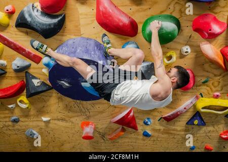 Vue latérale d'un homme fort dans des vêtements de sport grimpant en hauteur poignées murales colorées pendant les entraînements difficiles en salle de sport Banque D'Images