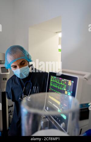 Vue latérale petit homme médecin dans l'uniforme médical regardant et vérifier dans un écran affichant l'équipement en fonctionnement léger chambre de l'hôpital contemporain Banque D'Images
