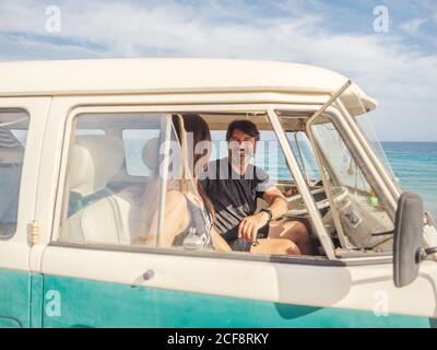 Homme barbu et femme aux cheveux longs dans des lunettes de soleil colorées assis devant la mer de la voiture et regardant la petite voiture bleue jouet Banque D'Images