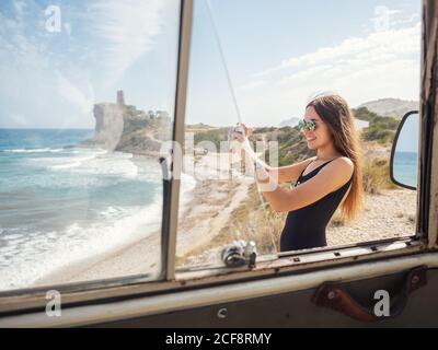 Jeune femme aux cheveux longs avec appareil photo en maillot de bain à la plage de sable avec vagues mousseuse Banque D'Images