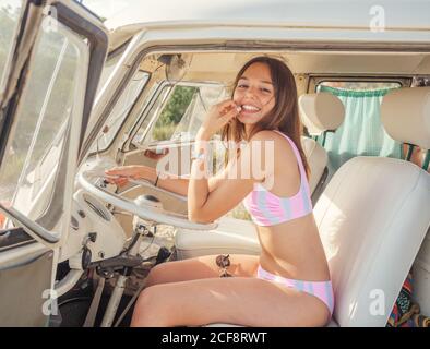 Jeune femme gaie souriant et regardant la caméra assis au volant de la voiture par beau temps ensoleillé Banque D'Images