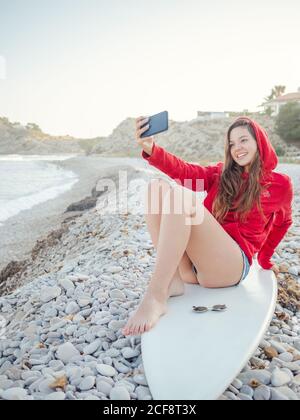 Jeune femme souriante à cheveux longs à capuche rouge prenant le selfie sur un smartphone allongé sur une planche de surf sur une côte pierreuse Banque D'Images