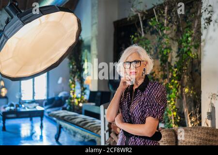 Actrice à cheveux gris pensive dans une position élégante à côté de la boîte souple et regarder la caméra pendant la pause dans le travail contre flou intérieur d'un studio contemporain confortable Banque D'Images