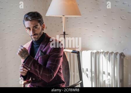 Homme adulte sérieux dans des vêtements élégants regardant l'appareil-photo pendant assis sur un fauteuil en bois ancien à côté d'un radiateur blanc et vintage lampadaire contre un mur blanc à pois dans la maison de campagne Banque D'Images