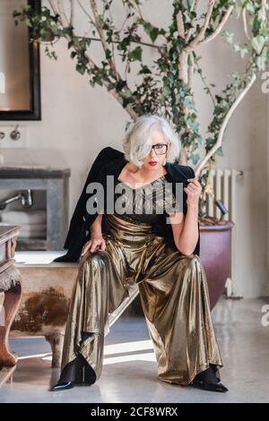 Calme senior confiante dame dans des lunettes et des vêtements élégants à la recherche à l'appareil photo tout en étant assis sur le bord de la baignoire de merde parmi les riches intérieur rétro avec meubles anciens et grand arbre à motif vert dans la chambre spacieuse de la maison de campagne Banque D'Images