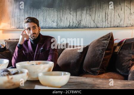 Homme pensif adulte dans des vêtements élégants regardant loin et méditer tout en étant assis sur un canapé doux en velours brun parmi les coussins à côté table en bois rustique avec grande image grise moderne dans spacieux appartement de style rétro Banque D'Images