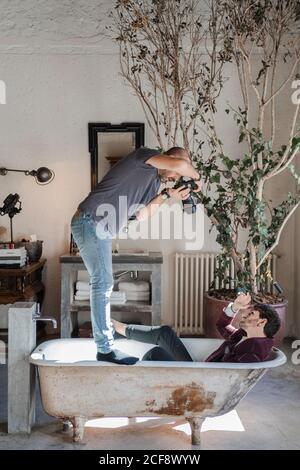 Vue latérale homme dédié debout sur les bords de la baignoire et de tournage d'en haut, un homme adulte dans des vêtements élégants couchés dans les vieux bain blanc près du grand arbre en pot vert contre rustique vintage intérieur en studio photo Banque D'Images