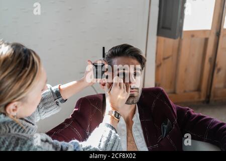 D'en haut diligent femme appliquant le maquillage sur l'homme barbu calme dans une élégante veste bordeaux assis dans un fauteuil avec un intérieur rustique flou dans un studio léger Banque D'Images