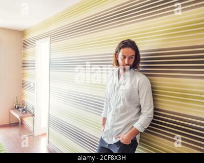 Homme barbu brutal dans une chemise décontractée et un Jean qui s'appuie sur mur à rayures dans l'appartement lumineux tenant les mains dans les poches regardant loin Banque D'Images