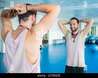 Jeune homme joyeux et barbu qui recueille les cheveux dans la queue en se tenant debout devant le miroir dans la salle de sport spacieuse Banque D'Images