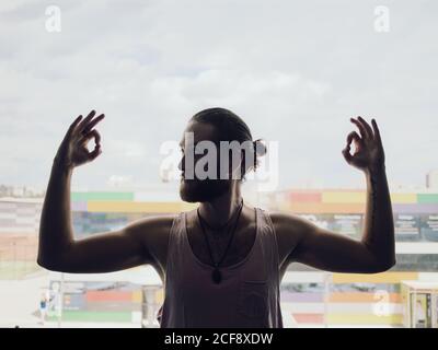 Jeune homme barbu dans des vêtements de sport effectuant l'exercice de yoga élevant mains et affiche ok près de la fenêtre avec vue de sity sur fond rétroéclairé Banque D'Images