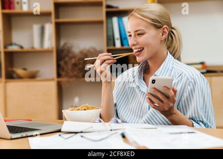 Image d'une jeune femme joyeuse mangeant des nouilles tout en travaillant avec ordinateur portable et téléphone portable au bureau Banque D'Images