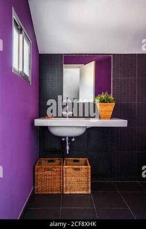 Salle de bains moderne avec mur violet coloré et grand miroir sur un élégant lavabo rectangulaire blanc avec paniers en paille en dessous Banque D'Images