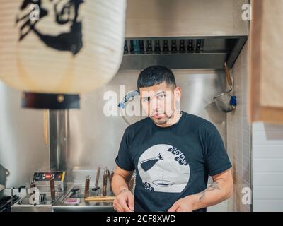Jeune homme ethnique qui cuisine un plat japonais et regarde la caméra Dans un café asiatique Banque D'Images