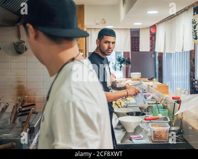 Vue latérale de jeunes hommes multiraciaux qui cuisent un plat japonais appelé Ramen dans le restaurant asiatique et regarder la caméra Banque D'Images