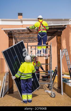 Groupe de travailleurs en uniforme et casques installant des panneaux photovoltaïques sur le toit de construction en bois près de la maison Banque D'Images