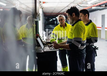 Groupe d'ingénieurs masculins qualifiés en uniforme utilisant des gadgets tout en examen de l'équipement électrique dans un bâtiment moderne Banque D'Images