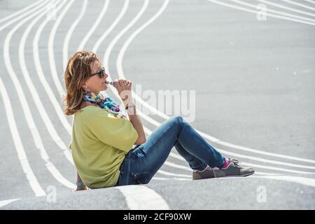 Vue latérale d'une femme qui fait froid chez un adulte dans des lunettes de soleil et un ensemble coloré qui exhalent de la vapeur tout en étant assise sur une route en asphalte tordant Banque D'Images