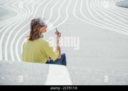 Vue latérale d'une femme qui fait froid chez un adulte dans des lunettes de soleil et un ensemble coloré qui exhalent de la vapeur tout en étant assise sur une route en asphalte tordant Banque D'Images