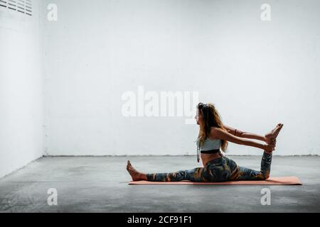 Vue latérale de la jeune femme flexible dans les vêtements de sport faisant devant s'asseoir sur le sol et se séparer avec la jambe relevée