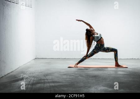 Vue latérale d'une femme calme portant des leggings et un soutien-gorge de sport debout sur le tapis en posture de guerrier inversé regardant vers le haut Banque D'Images