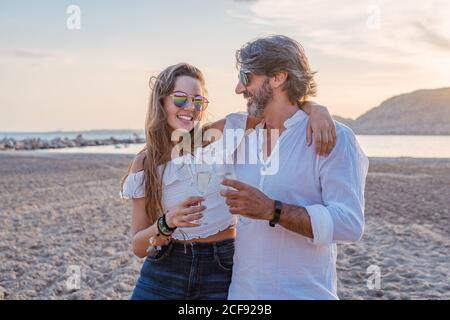 Homme mûr embrassant la jeune femme en joue tout en proposant des toasts et en célébrant la réunion de famille sur la plage de sable au crépuscule sur la station Banque D'Images