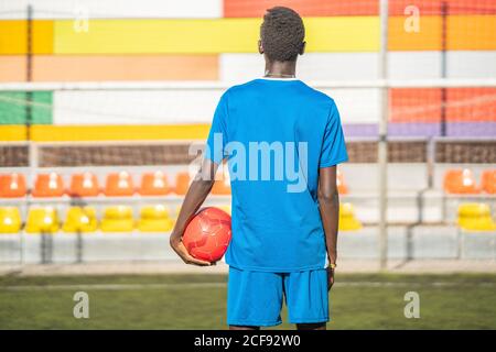 Vue arrière d'un homme noir dans un vêtement de sport bleu portant du rouge ballon en vous tenant contre les sièges du stade sur le terrain de football Banque D'Images