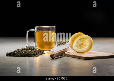 Boisson aromatique dans une tasse en verre avec des citrons et des tas de feuilles de thé séchées sur table sur fond noir Banque D'Images