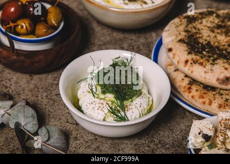 Bol de fromage recouvert de sauce verte servi pour le pain plat et olives sur table grise Banque D'Images