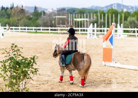 Vue arrière d'un enfant méconnaissable en costume de jockey assis selle d'un poney pendant la leçon dans l'école d'équitation Banque D'Images