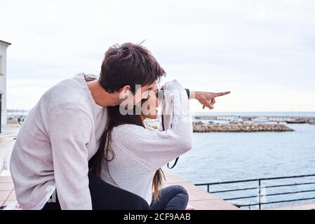 Vue latérale d'un couple romantique et gai dans une tenue décontractée assis sur la terrasse avec des jumelles et profiter de la vue tout en passant des vacances ensemble en bord de mer Banque D'Images