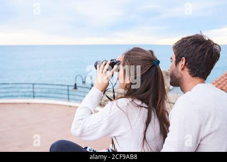 Vue latérale d'un couple romantique et gai dans une tenue décontractée assis sur la terrasse avec des jumelles et profiter de la vue tout en passant des vacances ensemble en bord de mer Banque D'Images