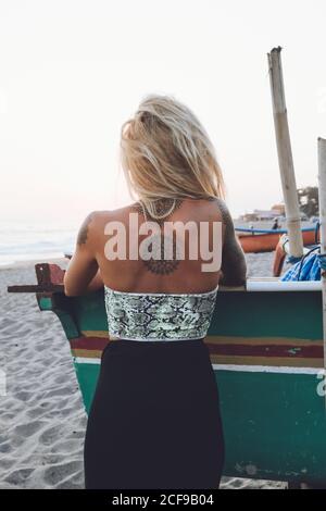 Vue arrière d'une jeune femme mince et méconnaissable en robe et avec le corps en tatouages se tenant sur la plage tropicale en admirant la mer en agitant au coucher du soleil Banque D'Images