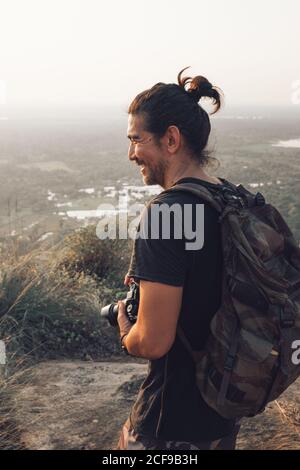 Vue latérale d'un jeune homme joyeux dans des vêtements décontractés avec une hersack debout sur le bord de la falaise et de prendre des photos de paysage majestueux de forêt verte au coucher du soleil Banque D'Images