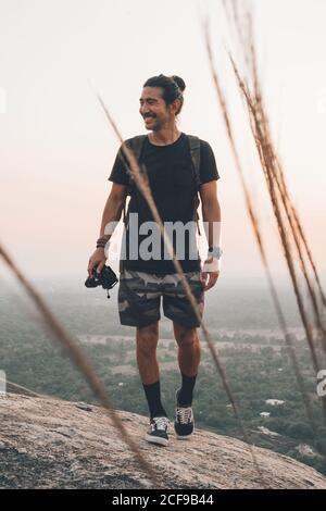 Jeune homme gai voyageur dans des vêtements décontractés avec photo debout au bord de la falaise et en prenant la photo de majestueux paysage de forêt verte au coucher du soleil Banque D'Images