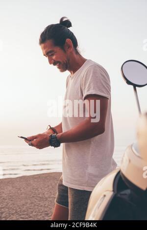 Vue latérale d'un homme sportif moderne positif dans un t-shirt et des courts-circuits à proximité du vélo et à l'aide d'un téléphone portable au bord de la mer au coucher du soleil Banque D'Images