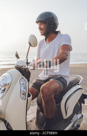 Vue latérale d'un joyeux homme actif habillé de blanc t chemise avec short et casque noir triporteur en été soirée sur la plage Banque D'Images