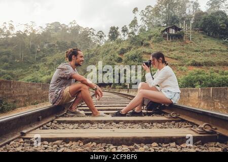 Vue latérale des jeunes hommes et femmes qui s'assoyent chemin de fer en face de l'autre et prendre des photos Appareil photo avec plantes vertes sur fond en Asie du Sri Lanka Banque D'Images