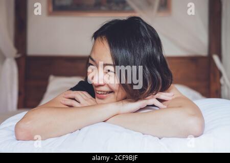 Femme asiatique optimiste regardant loin allongé sur un lit confortable et rire en étant allongé sur le lit le matin à la maison Banque D'Images