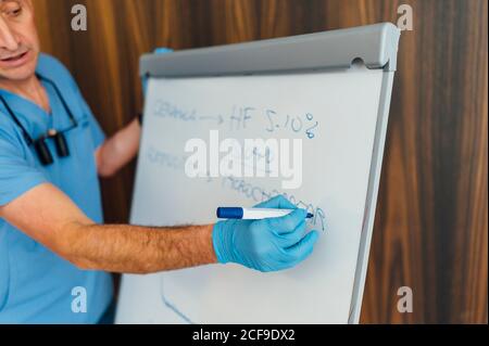 Vue latérale de la récolte mâle médecin en uniforme bleu et gants écrivant sur le tableau de conférence tout en expliquant la théorie pendant la leçon en cours de dentisterie Banque D'Images