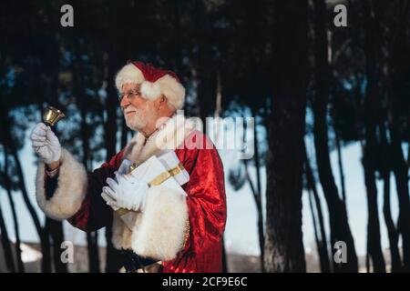 Homme senior gai en costume de Père Noël debout avec présenter et sonner dans les mains gantées en regardant loin sur la nature arrière-plan Banque D'Images