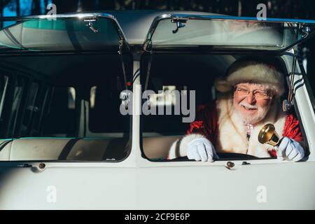 Homme senior souriant en costume de Père Noël en train de conduire vieux minibus avec cloche en métal à la main pour une journée ensoleillée loin Banque D'Images