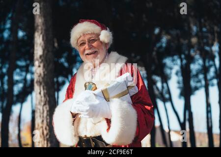 Homme senior gai en costume de Père Noël debout avec présenter et sonner les mains gantées en regardant l'appareil photo allumé arrière-plan de la nature Banque D'Images