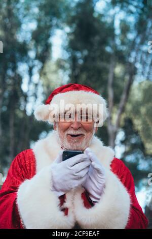 D'en-dessous, homme joyeux en costume de Père Noël en utilisant téléphone mobile moderne sur fond de nature floue Banque D'Images