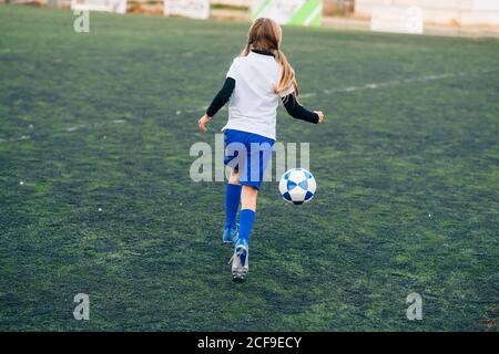 Vue arrière adolescente en uniforme blanc et bleu et chaussures de course à pied et préparation au ballon de football seul sur un terrain vert dans un club sportif contemporain Banque D'Images