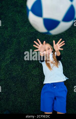 D'en haut joyeuse préteen fille en uniforme blanc et bleu lancer le ballon de football tout en étant couché seul sur le terrain vert dans club sportif moderne Banque D'Images