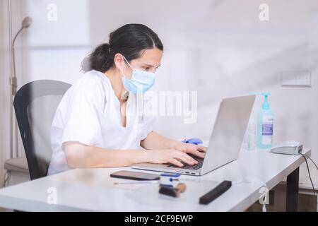 Jeune femme médecin sérieux portant un uniforme blanc et un masque médical travailler sur un ordinateur portable avec des gants en latex assis au bureau clinique moderne Banque D'Images